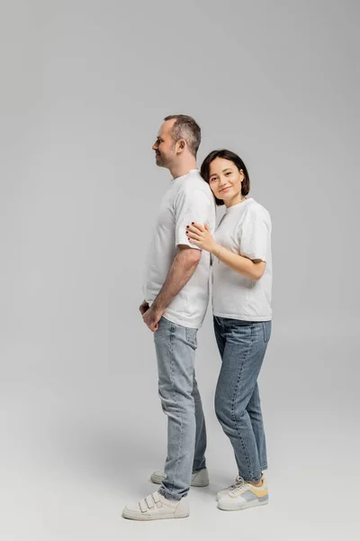Full length of tattooed woman with short brunette hair leaning on back of smiling husband while standing together in white t-shirts and denim jeans on grey background, happy couple — Stock Photo