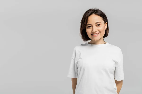 Happy and tattooed woman with short hair and natural makeup standing in white t-shirt and smiling while looking at camera isolated on grey background with copy space — Stock Photo