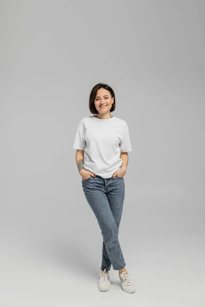 Full length of happy and tattooed woman with short hair and natural makeup standing in white t-shirt and posing with hands in pockets of blue denim jeans while looking at camera on grey background — Stock Photo
