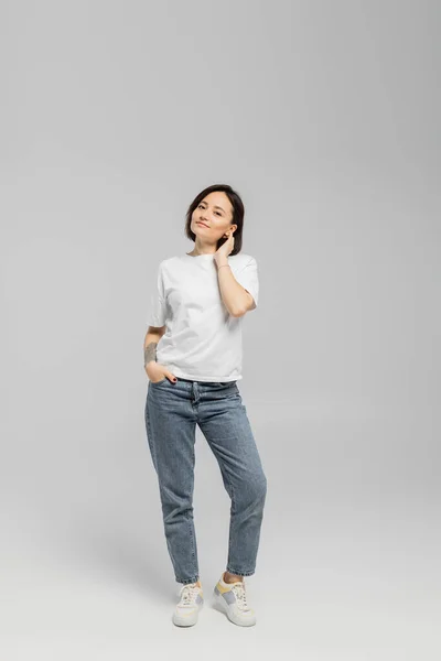 Full length of appealing and tattooed woman with short hair and natural makeup standing in white t-shirt and posing with hand in pocket of blue denim jeans while looking at camera on grey background — Stock Photo