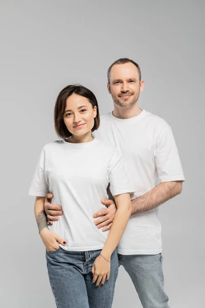 Cheerful husband hugging waist of tattooed wife with short brunette hair while standing together in white t-shirts and looking at camera isolated on grey background in studio, happy couple — Stock Photo