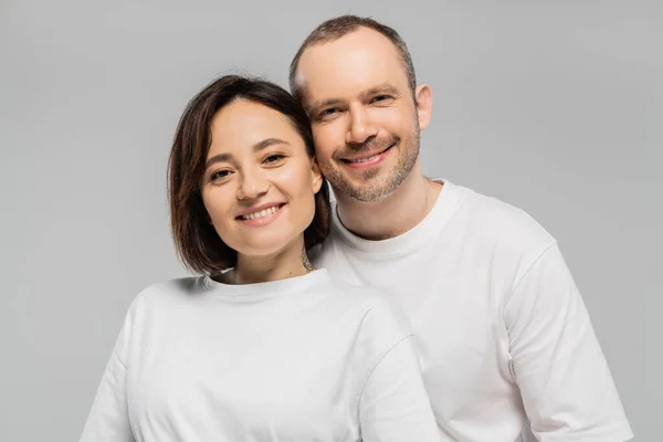 Marido alegre y esposa tatuada con pelo corto morena de pie juntos en camisetas blancas y mirando a la cámara aislada sobre fondo gris en el estudio, feliz pareja - foto de stock