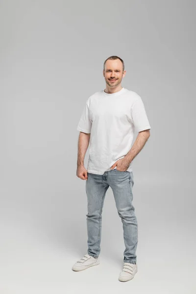 Full length of unshaved happy man with bristle standing in white t-shirt and posing with hand in pocket of blue denim jeans while looking at camera on grey background in studio, happiness — Stock Photo