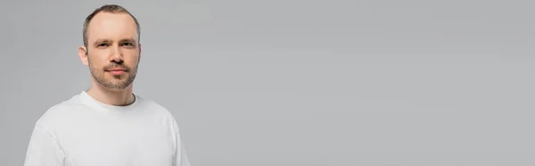 Hombre sin afeitar con cerdas de pie en camiseta blanca y mirando a la cámara mientras posa aislado sobre fondo gris en el estudio, espacio de copia, confianza y masculinidad, pancarta - foto de stock