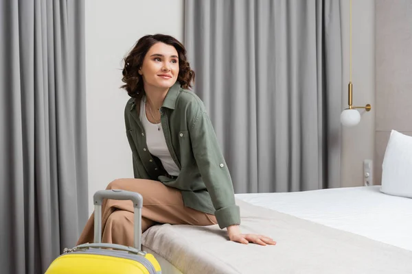 Молодая женщина с брюнетками, в повседневной рубашке и брюках, сидит на кровати рядом с желтым чемоданом, смотрит в сторону и улыбается в комфортабельном номере отеля, счастливый путешественник, выходные — стоковое фото