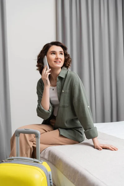 Weekend break, young and joyful woman with wavy brunette hair and tattoo, in casual clothes talking on mobile phone while sitting on bed near yellow suitcase and grey curtains in modern hotel suite — Stock Photo