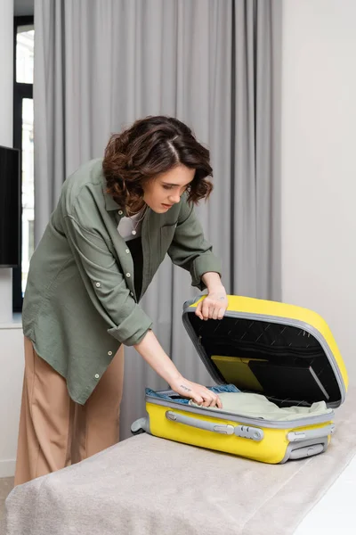 Weekend break, young and enchanting woman with wavy brunette hair and tattoo unpacking yellow suitcase with travel necessities near grey curtains, hotel experience, cozy atmosphere — Stock Photo