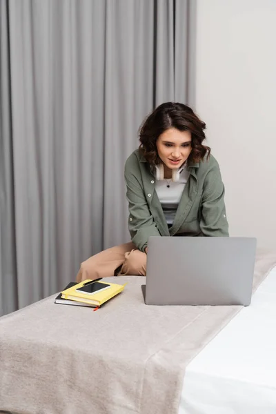 Цифровой кочевой образ жизни, улыбающаяся женщина с волнистыми волосами брюнетки и беспроводными наушниками, смотрящая на ноутбук, сидя рядом с ноутбуками, ручкой и смартфоном с чистым экраном на удобной кровати в номере отеля — стоковое фото