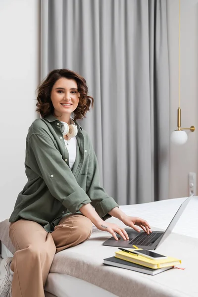 Joyful tattooed woman with wireless headphones sitting on bed and looking at camera near grey curtains, laptop, notebooks, pen and smartphone with blank screen in hotel, digital nomadism — Stock Photo
