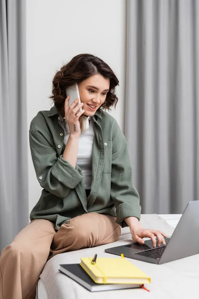 Cheerful tattooed woman with wireless headphones talking on mobile phone and working on laptop while sitting near grey curtains, notebooks and pen on bed in comfortable hotel room, phone call — Stock Photo