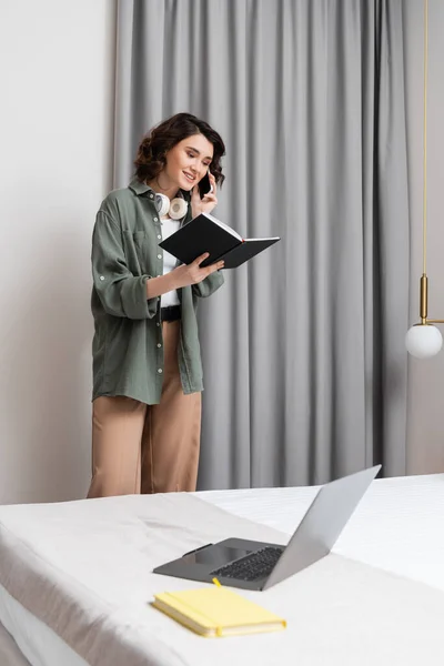 Estilo de vida independiente, mujer feliz con auriculares inalámbricos mirando el cuaderno y hablando en el teléfono móvil cerca de cortinas grises, aplique de pared, ordenador portátil y bloc de notas en la cama cómoda en la habitación de hotel moderna - foto de stock