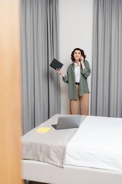 Freiberuflicher Lebensstil, fröhliche junge Frau in lässiger Kleidung mit kabellosen Kopfhörern und Notebook, während sie neben grauen Vorhängen und Bett mit Notizblock und Laptop im Hotelzimmer steht — Stockfoto