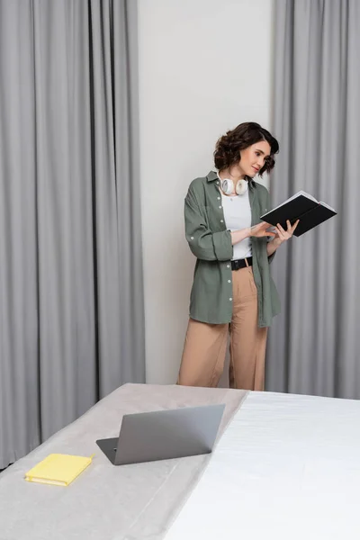 Freelancer, young woman with wavy brunette hair and wireless headphones looking at notebook while standing in casual clothes near grey curtains, laptop and notepad on bed in modern hotel suite — Stock Photo