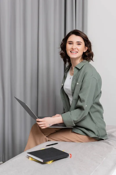 Carefree woman with wavy brunette hair sitting on bed with laptop and looking at camera near grey curtains, pen and notebooks in hotel room, work and travel, freelance lifestyle — Stock Photo