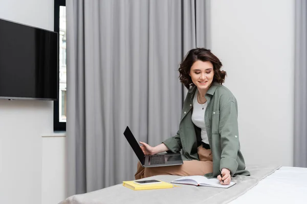 Remote work, smiling woman with wavy hair writing in notebook and looking at camera while sitting near grey curtains, notepad and smartphone with blank screen on bed in modern hotel room — Stock Photo