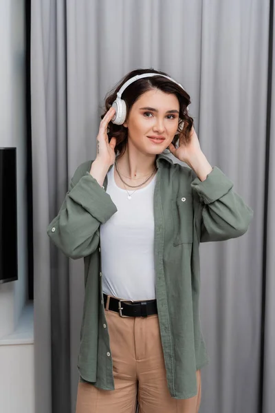 Pleased woman with wavy brunette hair and tattoo looking at camera while listening podcast in wireless headphones near grey curtains in modern hotel suite, leisure, weekend getaway — Stock Photo