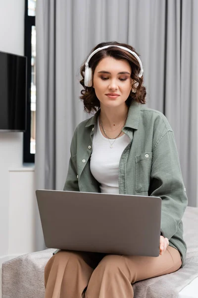 Young woman with wavy brunette hair, casual clothes and wireless headphones sitting on bed with laptop during educational webinar near grey curtains in hotel suite, study and travel — Stock Photo