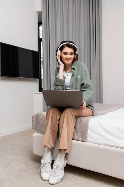 Happy young woman with wavy brunette hair and tattoo sitting on bed with laptop near lcd tv, grey curtains, adjusting wireless headphones during educational webinar in hotel suite, study and travel — Stock Photo