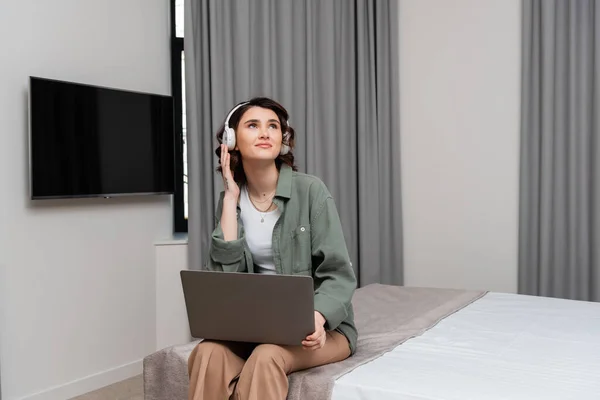 Позитивная молодая женщина регулируя беспроводные наушники и глядя в сторону, сидя на кровати с ноутбуком возле ЖК-телевизор и серые шторы во время онлайн-урока в номере отеля, учебы и путешествий — стоковое фото