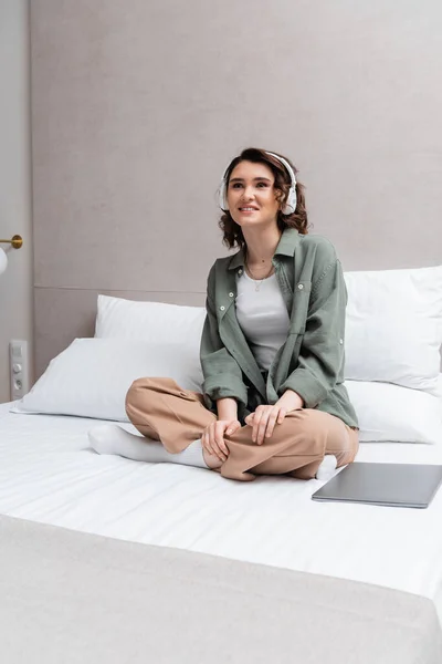 Joyful woman with brunette hair, in casual clothes and headphones sitting on bed with crossed legs and listening musical podcast in wireless headphones near white pillows and laptop in hotel room — Stock Photo