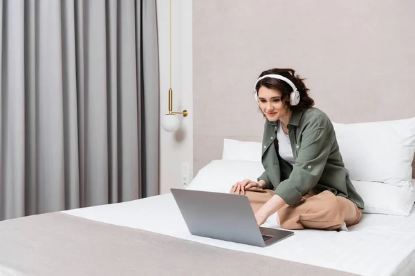 Mujer joven feliz en ropa casual y auriculares inalámbricos sentados en la cama cerca de almohadas blancas, cortinas grises y aplique de pared mientras ve la película en el ordenador portátil en la habitación de hotel, ocio y viajes - foto de stock