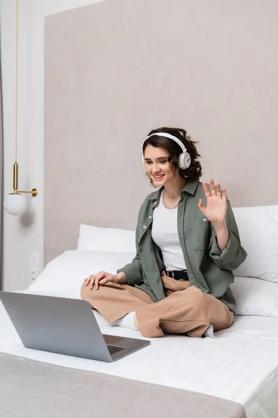 Overjoyed woman with wavy brunette hair, in wireless headphones and casual clothes sitting on bed and waving hand during video call on laptop near white pillows and wall sconce in hotel room — Stock Photo