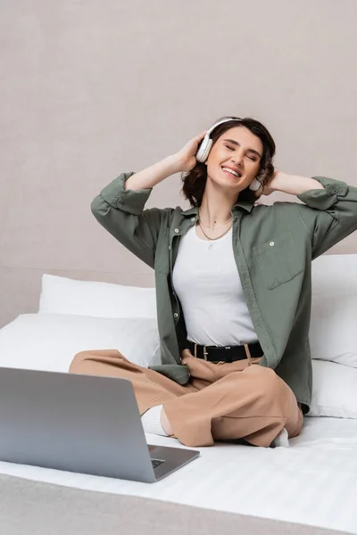 Carefree woman with wavy brunette hair and closed eyes listening musical podcast in wireless headphones while sitting on bed near laptop and white pillows in hotel suite, leisure and travel — Stock Photo