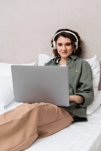 Carefree woman with wavy brunette hair sitting on bed near white pillows in wireless headphones and casual clothes and watching film on laptop in comfortable hotel room, leisure and travel — Stock Photo