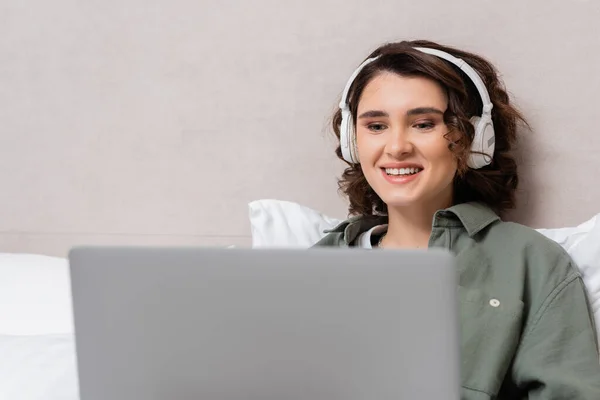 Mujer joven y alegre con el pelo ondulado morena viendo la película en el ordenador portátil borroso mientras está sentado en los auriculares inalámbricos cerca de la pared gris y almohadas blancas en la habitación de hotel moderna - foto de stock