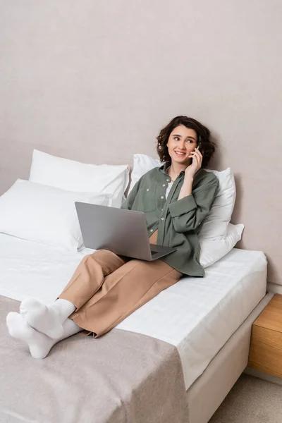 Full length of young and happy woman in casual clothes, with wavy brunette hair looking away while sitting on bed with laptop and talking on mobile phone near white pillows in hotel suite — Stock Photo