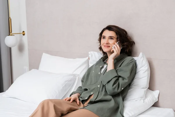 Pleased young woman in casual clothes, with wavy brunette hair talking on mobile phone near grey wall and white pillows on comfortable bed in model hotel room, leisure and travel — Stock Photo