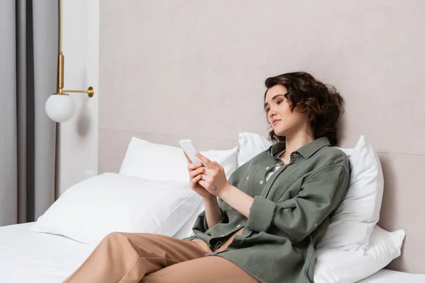 Mujer joven con el pelo ondulado morena y tatuaje sentado en la cama en Internet cerrado casual y navegación en el teléfono móvil cerca de almohadas blancas y aplique de pared en la habitación de hotel cómodo, ocio y viajes - foto de stock