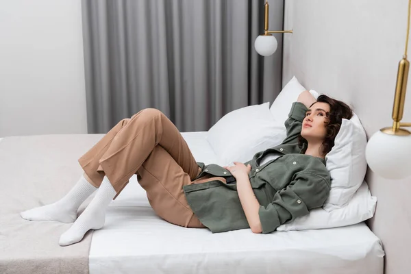 Full length of dreamy and carefree woman with wavy brunette hair laying on bed in casual clothes near white pillows, wall sconces and grey curtains in cozy atmosphere of modern hotel room — Stock Photo
