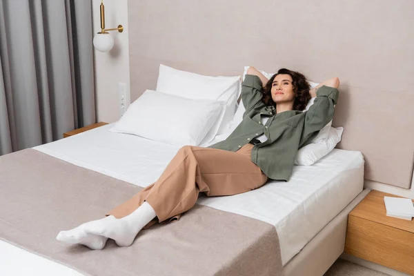 Full length of young delighted woman with wavy brunette hair lying on bed and white pillows in casual clothes near bedside table, books and wall sconce in contemporary hotel room — Stock Photo