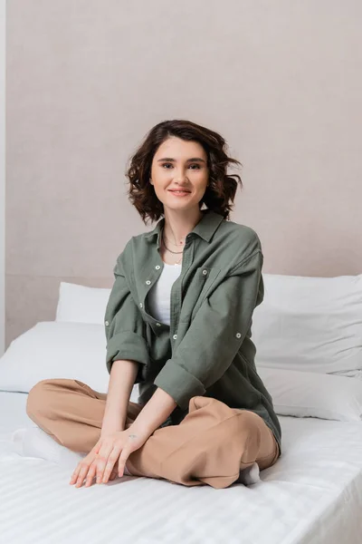 Carefree woman with wavy brunette hair sitting on bed in casual clothes, with crossed legs, looking at camera near white pillows and grey wall in contemporary hotel suite, leisure and travel — Stock Photo
