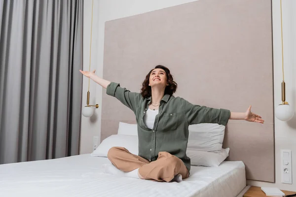 Young excited woman with wavy brunette hair and in casual clothes sitting on bed with outstretched hands and looking up near white pillows, wall sconces and grey curtains in comfortable hotel room — Stock Photo