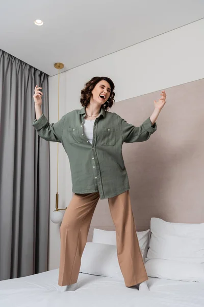 Young excited woman with wavy brunette hair, wearing casual clothes, dancing and singing on bed near white pillows, wall sconce and grey curtains in cozy hotel suite — Stock Photo