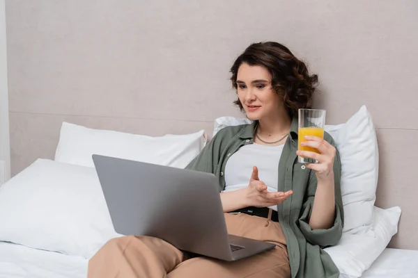 Mujer sonriente con cabello castaño ondulado sosteniendo un vaso de jugo de naranja fresco y un gesto durante la videollamada en el portátil mientras está sentada en la cama cerca de almohadas blancas y pared gris en la habitación de hotel - foto de stock