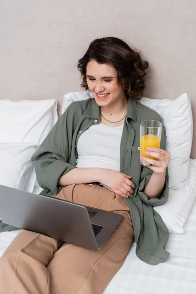 Young and carefree woman in casual clothes, with wavy brunette hair and tattoo holding glass of fresh orange juice while watching movie on laptop on bed near white pillows and grey wall in hotel room — Stock Photo