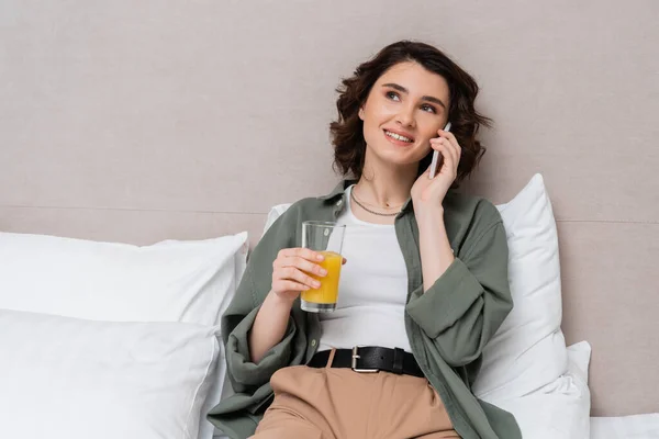 Phone call, young carefree woman holding glass of fresh orange juice and talking on smartphone while sitting on bed in casual clothes near white pillows and grey wall in hotel room — Stock Photo