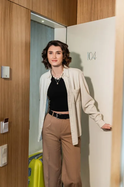 Delighted woman in stylish clothes, white shirt and beige pants, with wavy brunette hair, holding yellow suitcase and opening door of modern hotel room with secure entry and keyboard access — Stock Photo