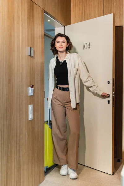 Full length of pleased woman in white shirt and beige pants, with wavy brunette hair holding yellow travel bag and smiling while opening door of hotel suite near keycard reader, check-in — Stock Photo