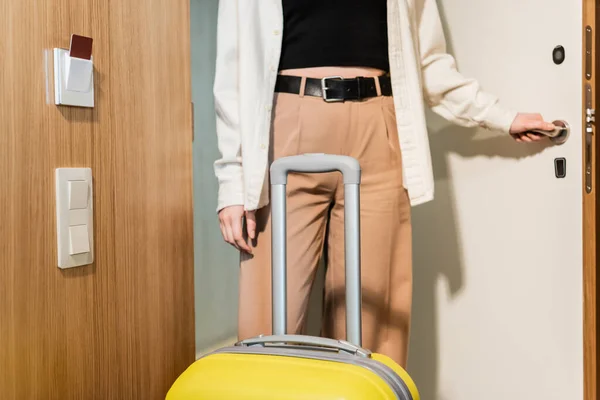 Обрезанный вид молодой и стильной женщины в белой рубашке и бежевых брюках, открывающих входную дверь в современном отеле рядом с желтой дорожной сумкой и считывателем карт-ключей, безопасный вход, стиль жизни в путешествии, регистрация заезда — стоковое фото