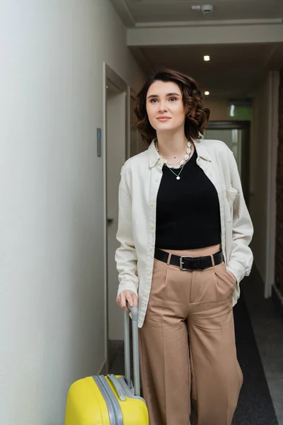 Young woman with wavy brunette hair, in black crop top, white shirt and beige pants walking with hand in pocket and yellow suitcase along corridor in contemporary hotel — Stock Photo