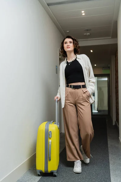 Mujer de moda con el pelo ondulado morena, con camisa blanca, top negro y pantalones beige de la mano en el bolsillo mientras camina con la maleta amarilla a lo largo del pasillo en el hotel moderno - foto de stock