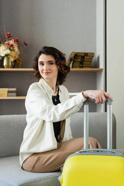 Carefree tattooed woman with wavy brunette hair, in stylish casual clothes looking at camera while sitting on couch with yellow suitcase, near shelves with books and vase with flowers in hotel lobby — Stock Photo