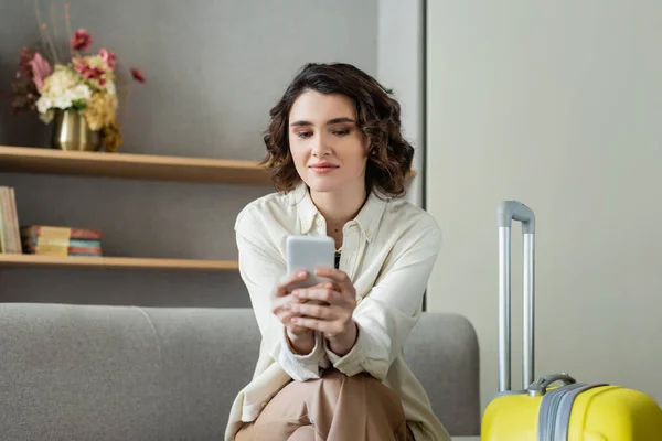 Young smiling woman with brunette hair using smartphone while sitting on couch near travel bag, shelves with books and vase with flowers on blurred background in hotel lobby, social networking — Stock Photo