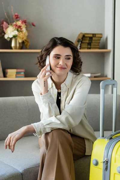 Happy young woman with wavy brunette hair and tattoo sitting on couch and talking on mobile phone near yellow travel bag, books and vase with flowers on shelves in hotel lobby on blurred background — Stock Photo