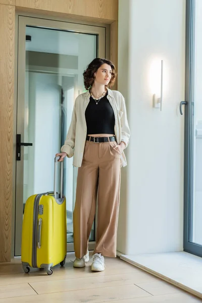 Full length of young and confident woman in white shirt, black crop top and beige pants standing with hand in pocket near yellow travel bag and glass door in lobby of contemporary hotel — Stock Photo