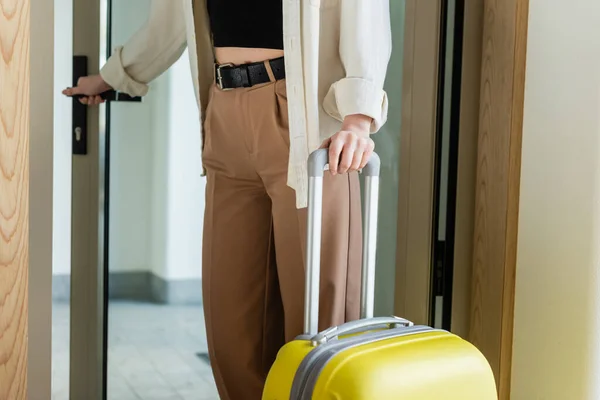Vista parcial de la mujer joven y de moda en camisa blanca, top negro, pantalones beige y cinturón de cuero de pie con bolsa de viaje amarilla cerca de la puerta de cristal al entrar en el vestíbulo del hotel moderno - foto de stock